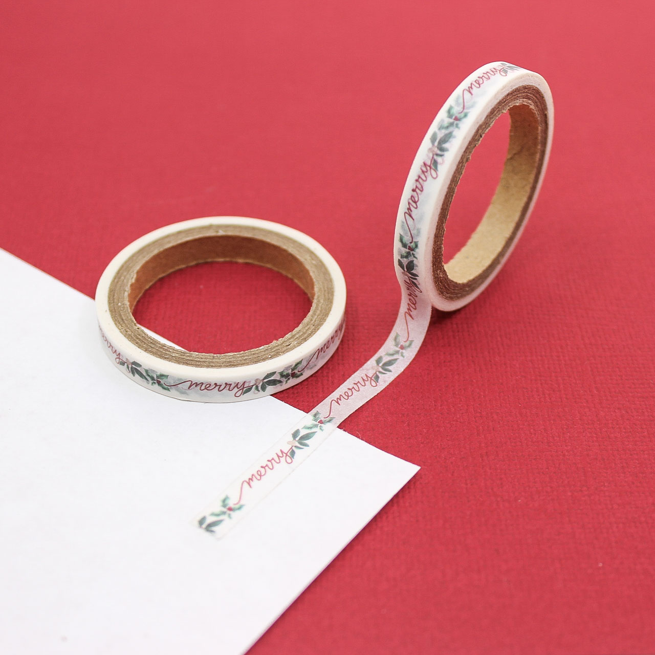 Narrow holly and merry script washi tape roll displayed on red background.