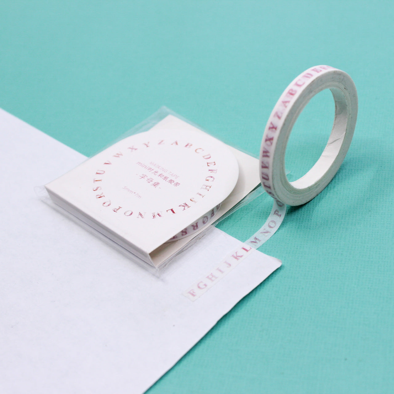 Narrow washi tape with pink alphabet letters rolled out on white paper against mint background.