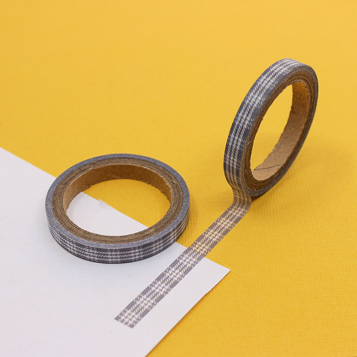 Narrow grey plaid washi tape roll on yellow background with strip applied to paper.
