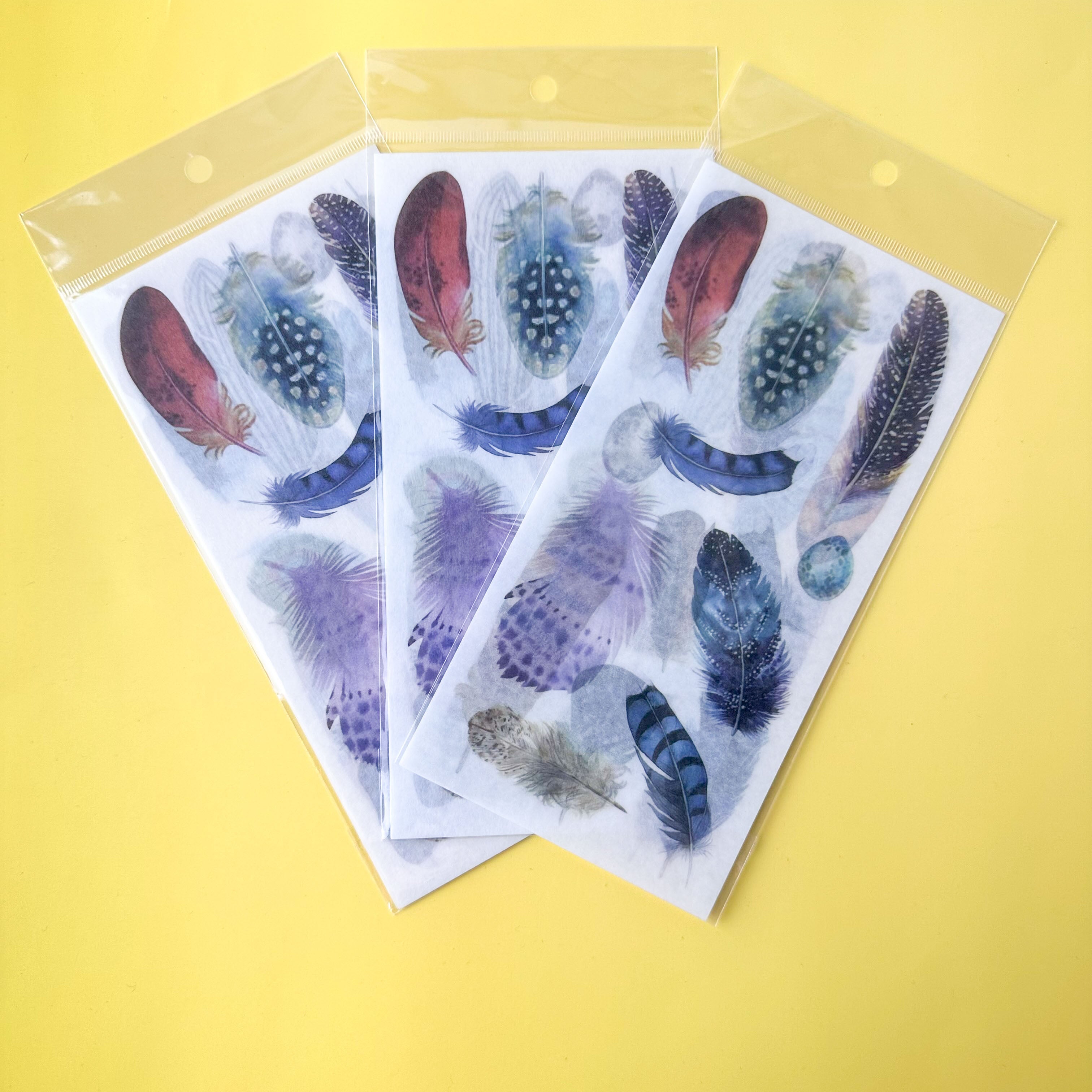 Three sheets of feather-themed stickers in clear plastic packaging on a yellow background