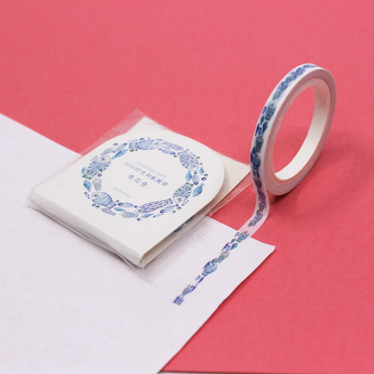 Narrow washi tape with watercolor blue fish pattern unrolled onto white paper on a pink background.