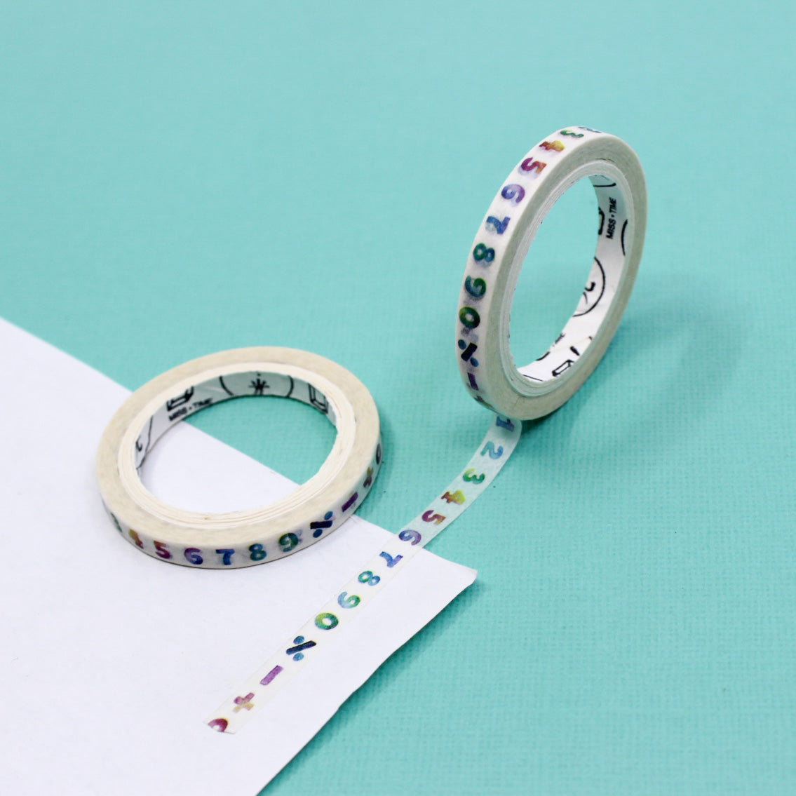 Narrow washi tape roll with rainbow-colored numbers unrolled across white planner paper.