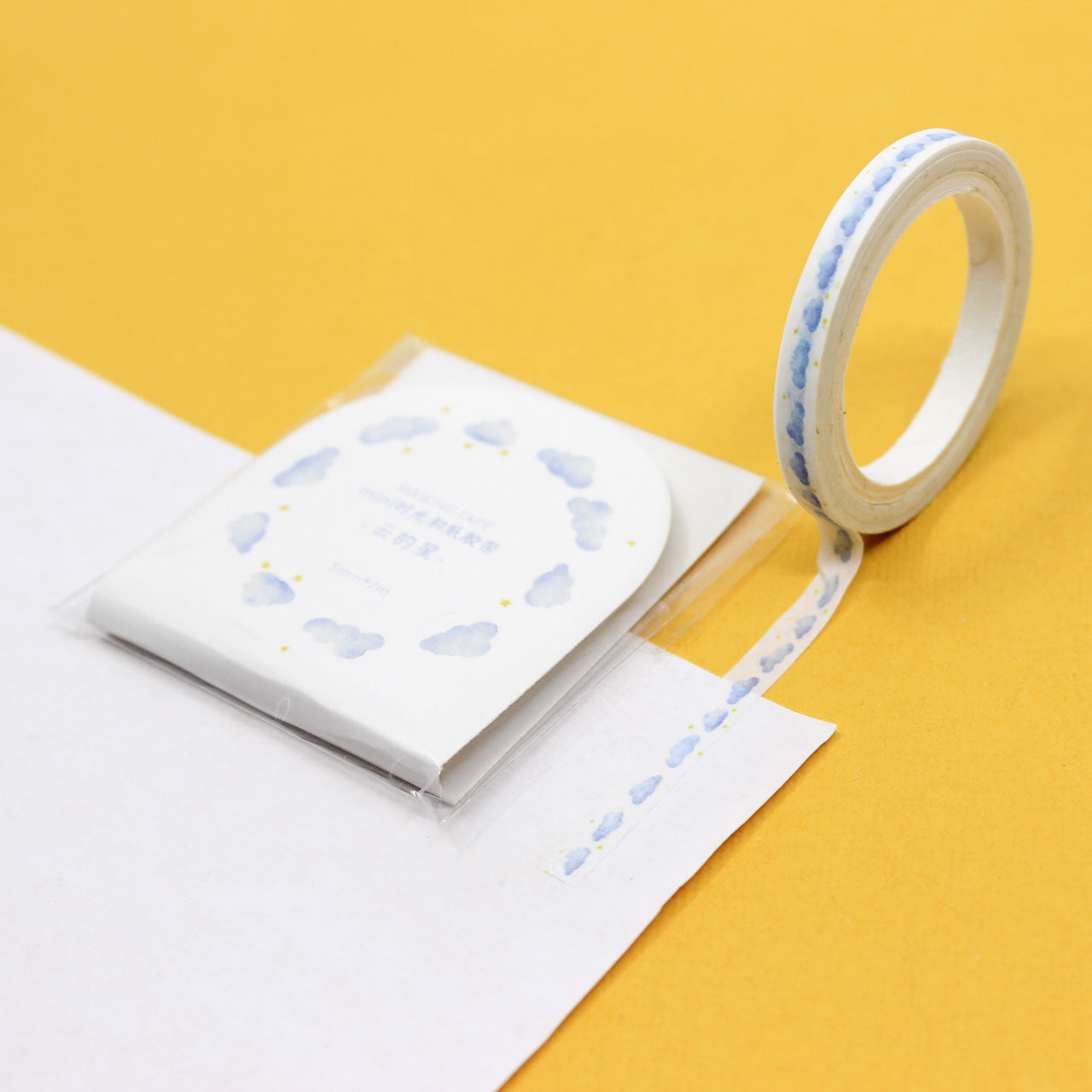 Roll of narrow washi tape decorated with small watercolor blue clouds on a yellow background.