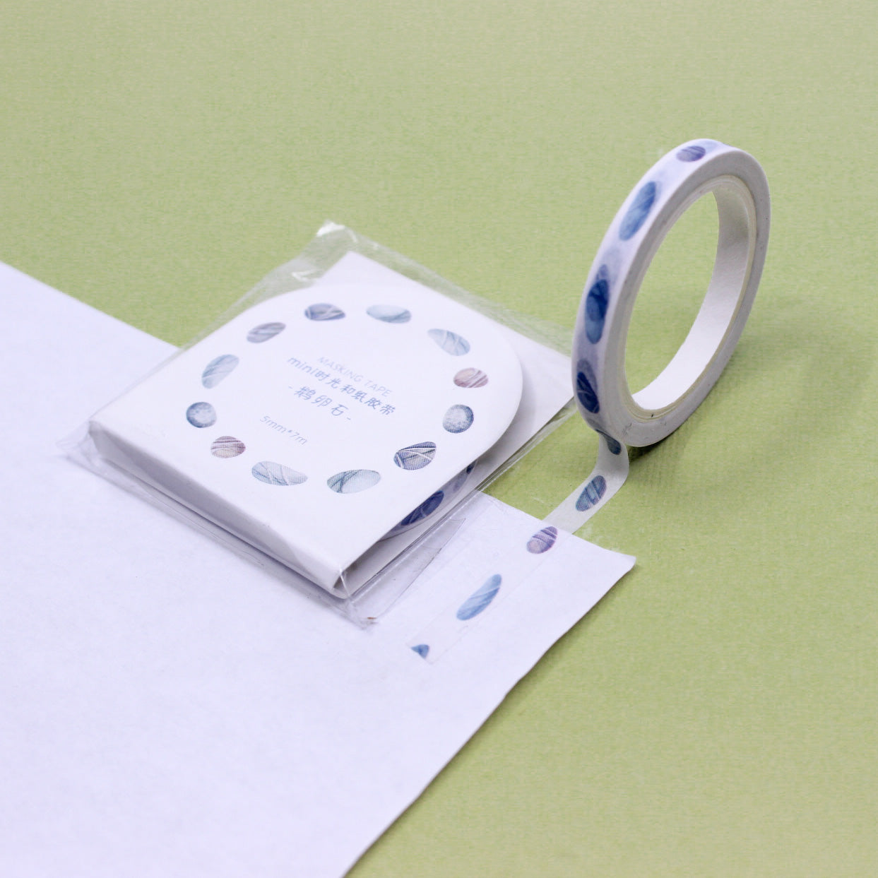 Narrow washi tape with watercolor pebble pattern rolled out on white paper against a light green background.